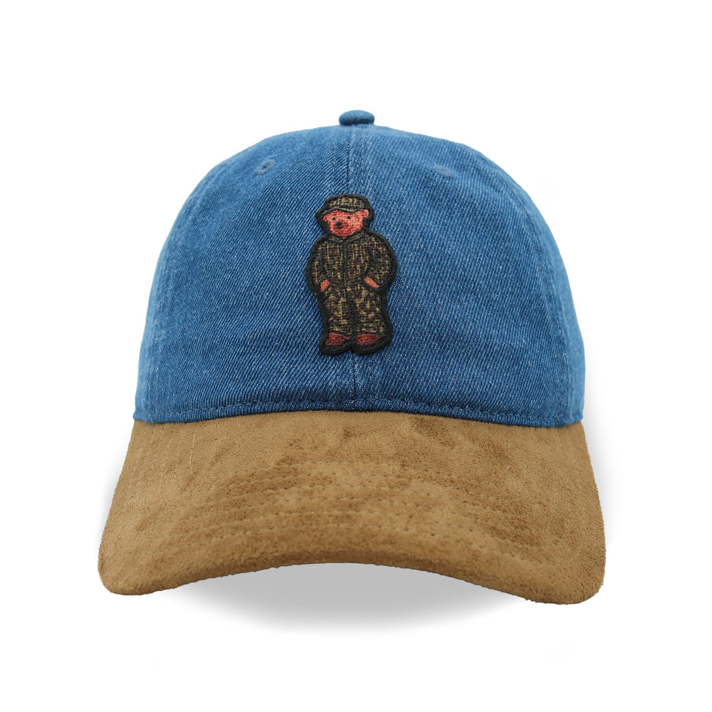 A baseball cap with a blue denim crown and a brown suede brim, featuring an embroidered teddy bear patch on the front.
