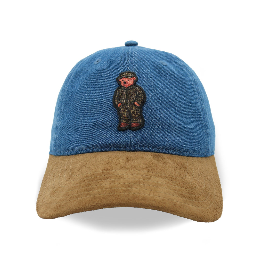 A baseball cap with a blue denim crown and a brown suede brim, featuring an embroidered teddy bear patch on the front.