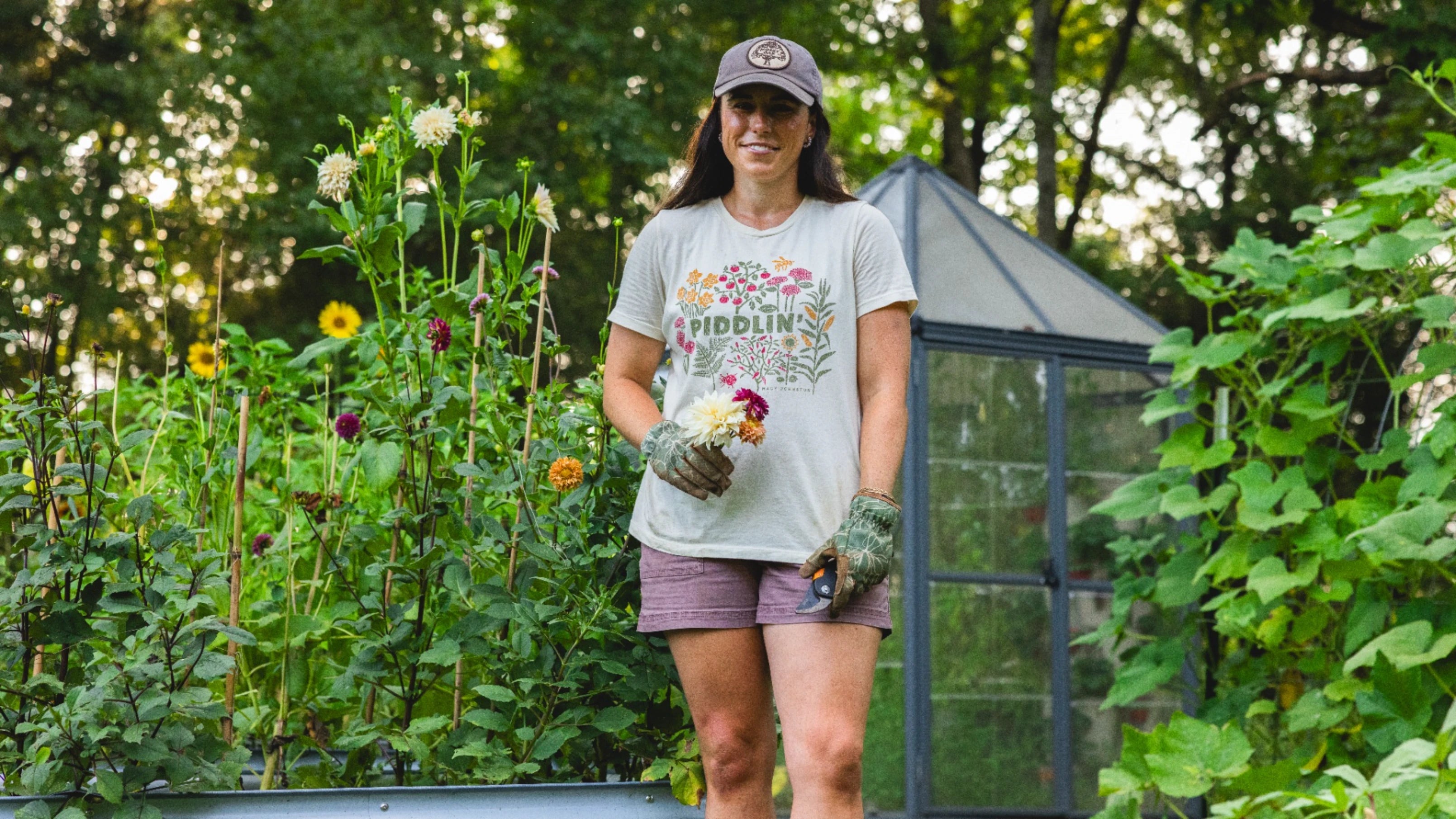 A woman stands in a garden wearing a gray graphic T-shirt that reads 'FIDDLIN'', purple shorts, gardening gloves, and a cap, with a greenhouse and leafy plants in the background.