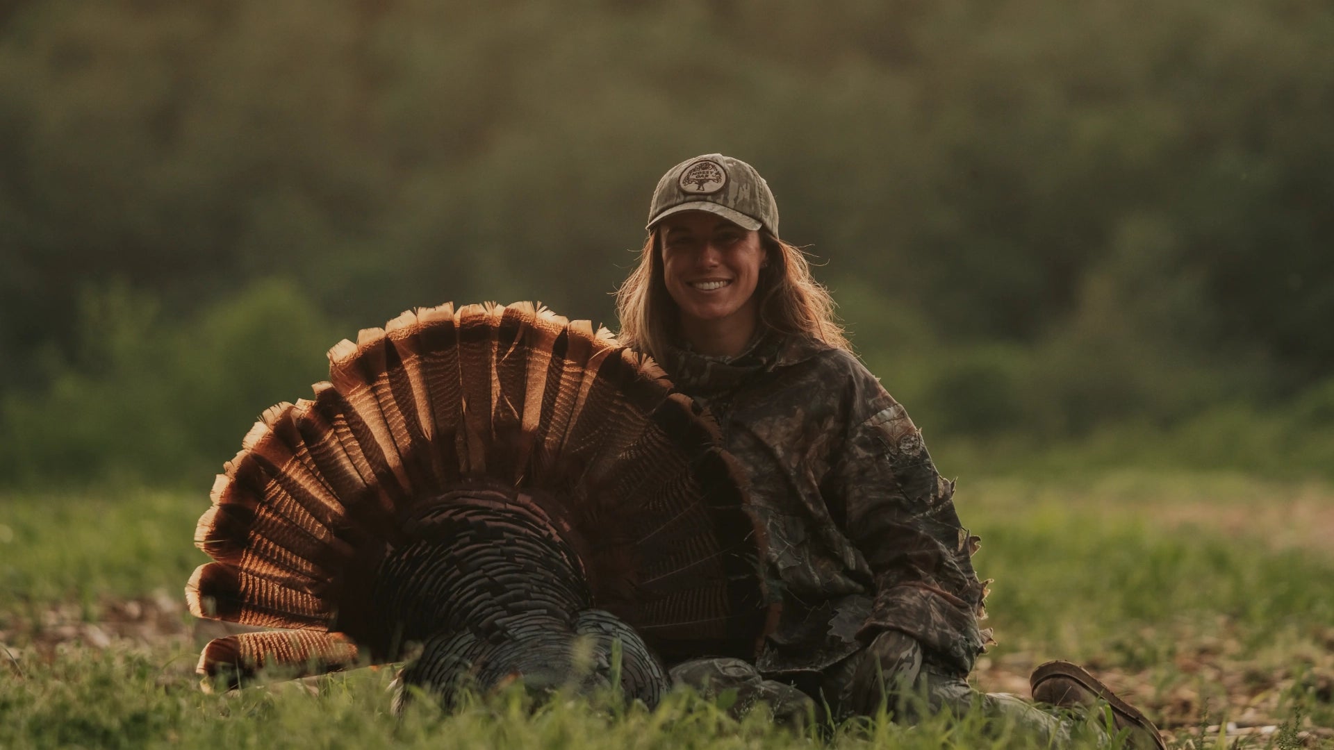 Macy in camouflage clothing and a cap sits in a grassy outdoor field with a large wild turkey in front of her, displaying a fanned tail.