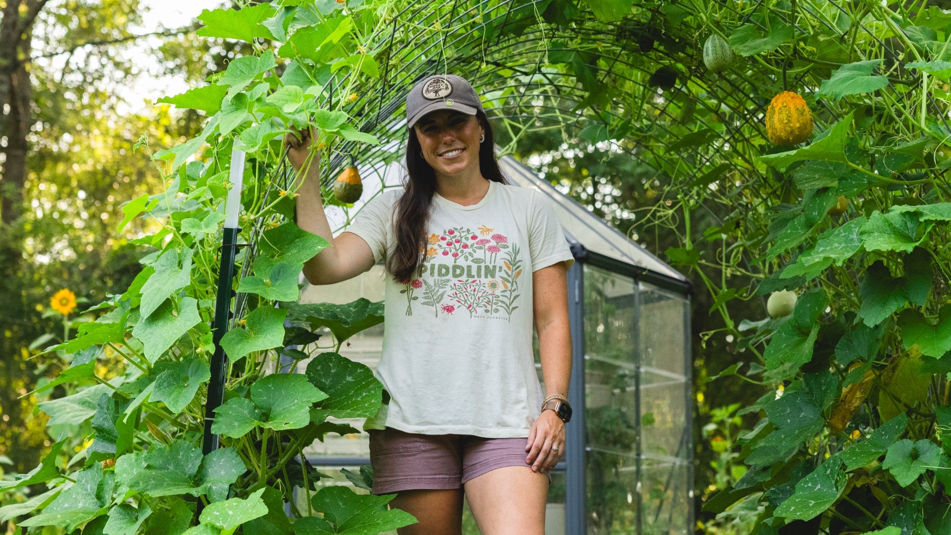 A woman stands in a lush garden greenhouse wearing a white graphic t-shirt with a colorful floral design and the word 'PIDDLIN'', paired with mauve-purple shorts. She is surrounded by green cucumber vines, hanging melons, and a small glass greenhouse structure.