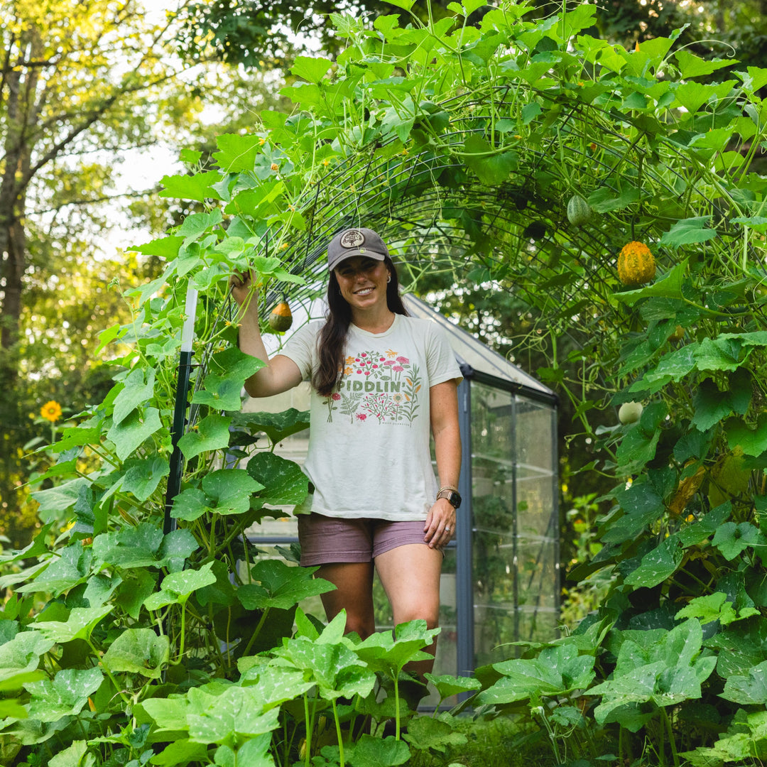 Macy W Johnston stands in a lush garden greenhouse wearing a white graphic t-shirt that reads 'Piddlin', purple shorts, and a baseball cap, surrounded by green vines and small yellow-orange gourds.