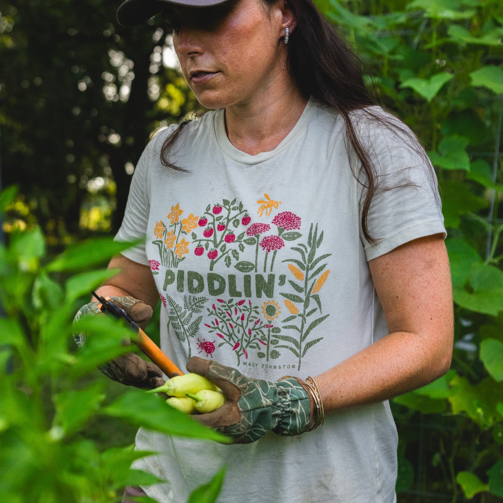 Macy W Johnston wearing as sage t-shirt with a colorful floral design and the text 'PIDDLIN'' while gardening in a lush green outdoor setting.