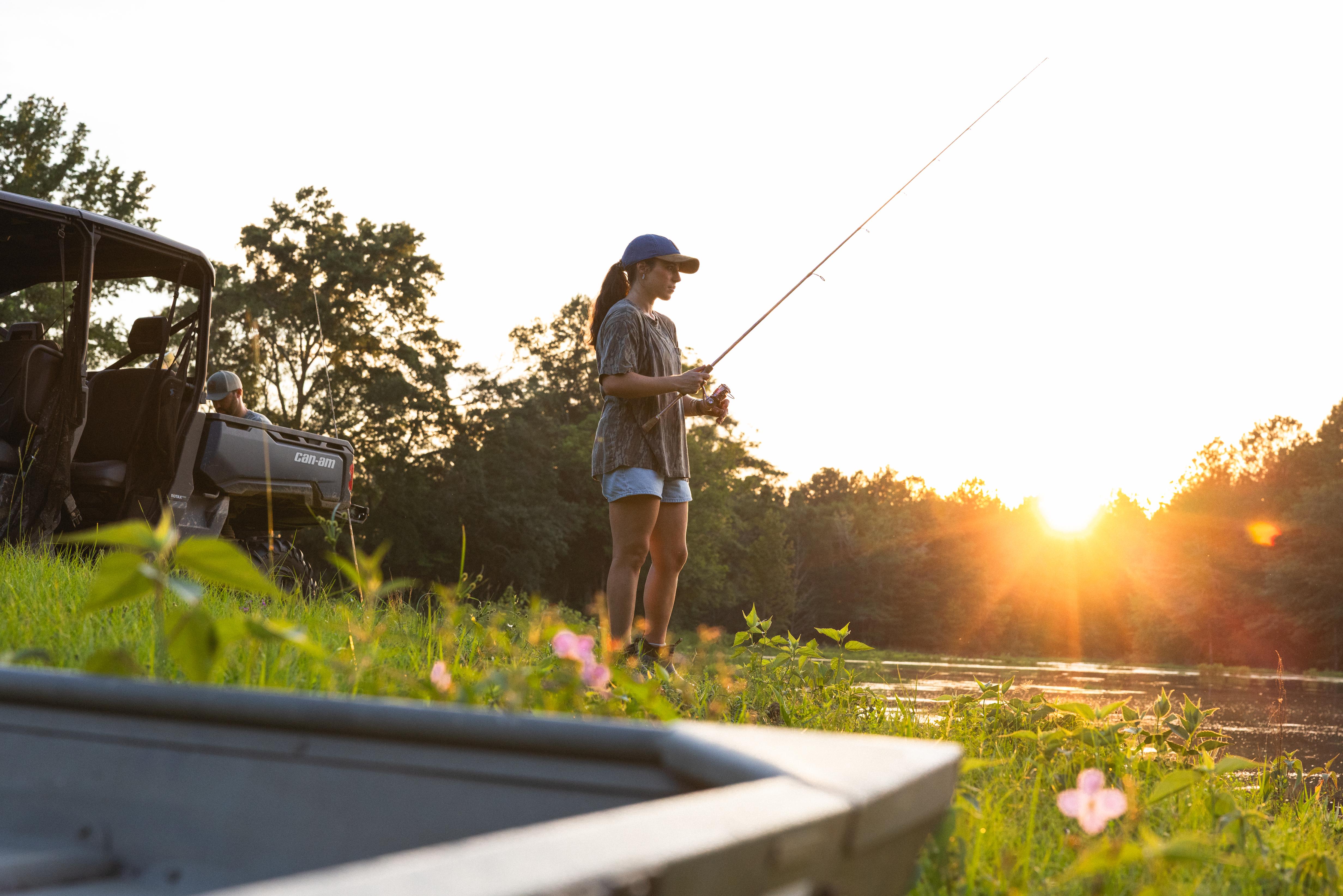 A woman stands in a grassy outdoor area at sunset, holding a fishing rod near a pond or river, with trees in the background and acan-am on the left.