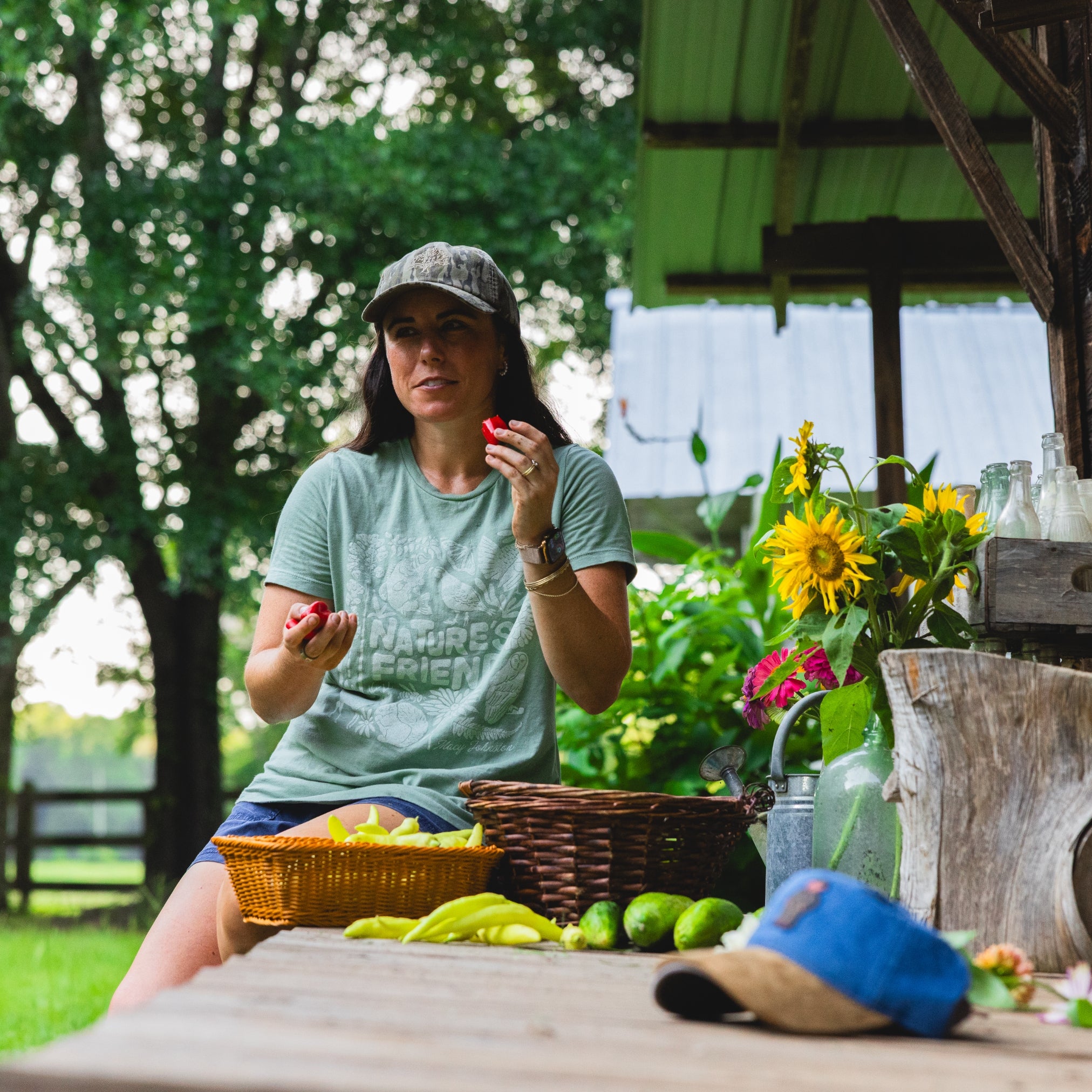 Macy Johnston wearing a light green graphic t-shirt and a gray baseball cap sits at a wooden table outdoors, holding small red fruits. In front of her are baskets, yellow flowers, and limes on a rustic table under a green-roofed shelter.