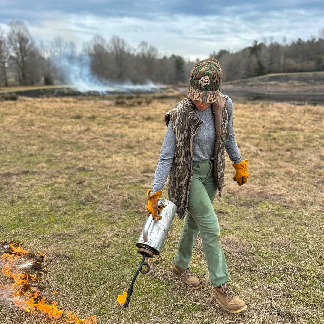 A person standing in an open field wearing a camouflage cap, gray long-sleeve shirt, brown vest, green pants, and tan boots, while holding a metal propane torch with a flame nozzle. Smoke rises in the background, and dry grass with a small fire is visible in the foreground.
