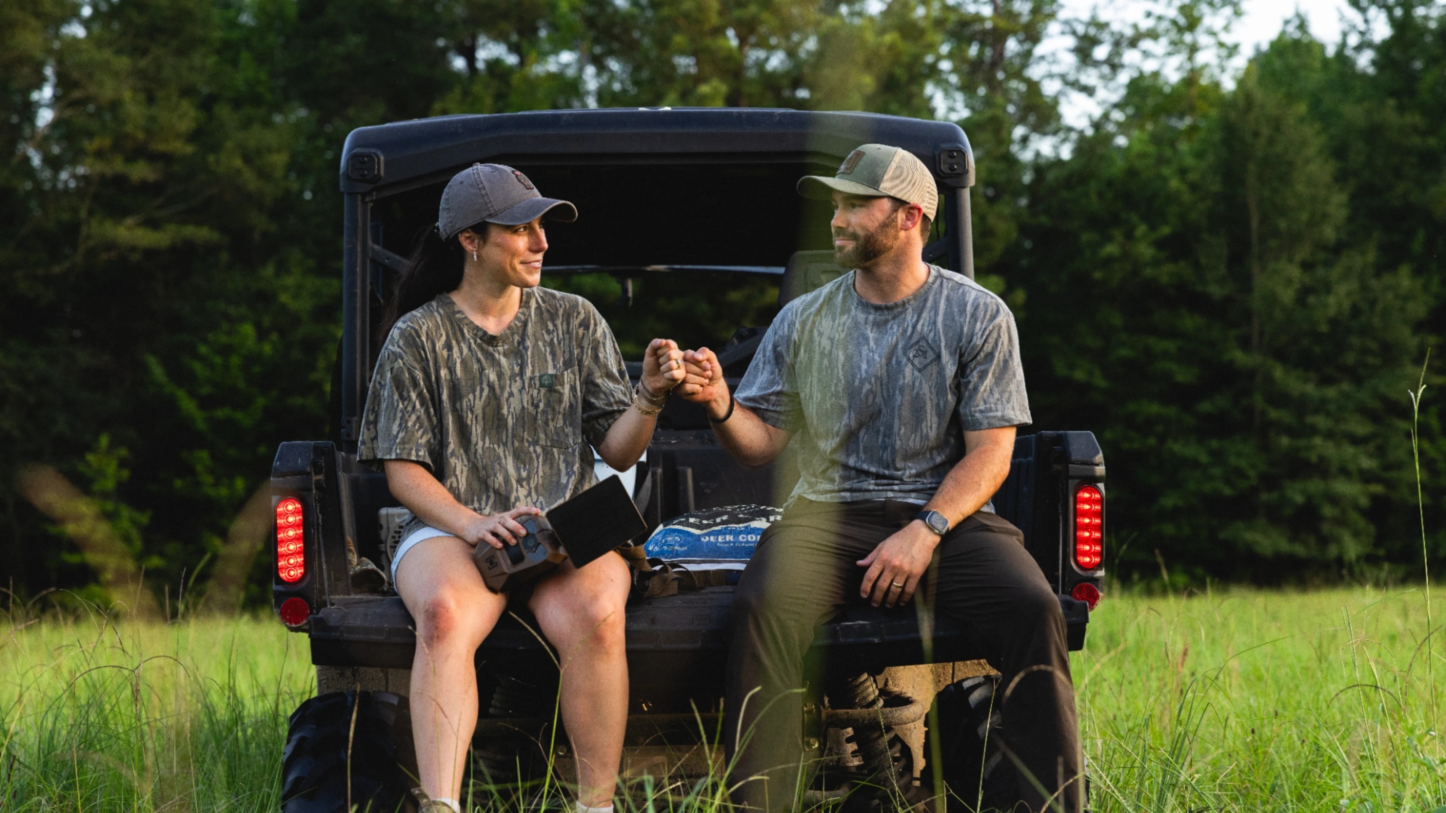 Macy and Slade Johnston sitting on the back of a black utility vehicle in a grassy field, wearing matching camouflage t-shirts and caps while holding hands.
